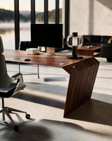 EDELHOLZ DeskPURE Schreibtisch in Deep Walnut, Raumansicht mit Bürostuhl, Monitor und Tischleuchte vor bodentiefen Fenstern [finish:deep-walnut]